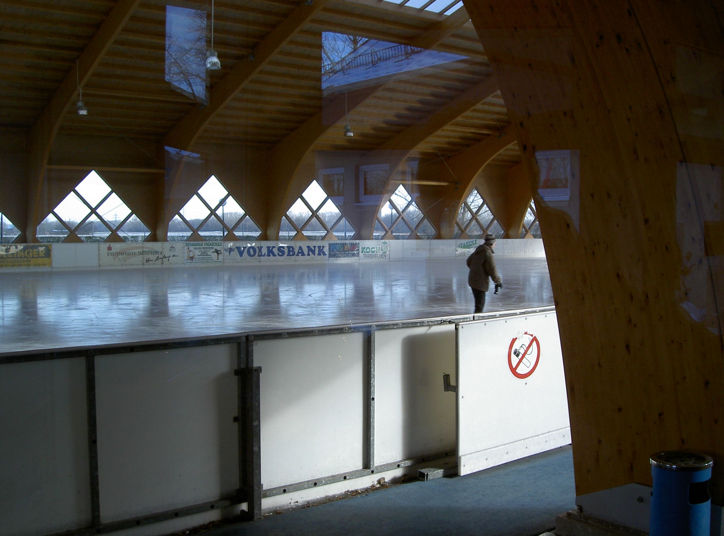 Egy fedett műjégpálya, amelyen egy ember korcsolyázik. A pályát fagerendák és ablakok veszik körül.