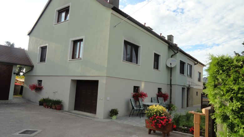 Egy k&eacute;tszintes h&aacute;z k&uuml;lső n&eacute;zete, előt&eacute;rben z&ouml;ld fest&eacute;kkel, vir&aacute;gokkal &eacute;s kerti b&uacute;torokkal.