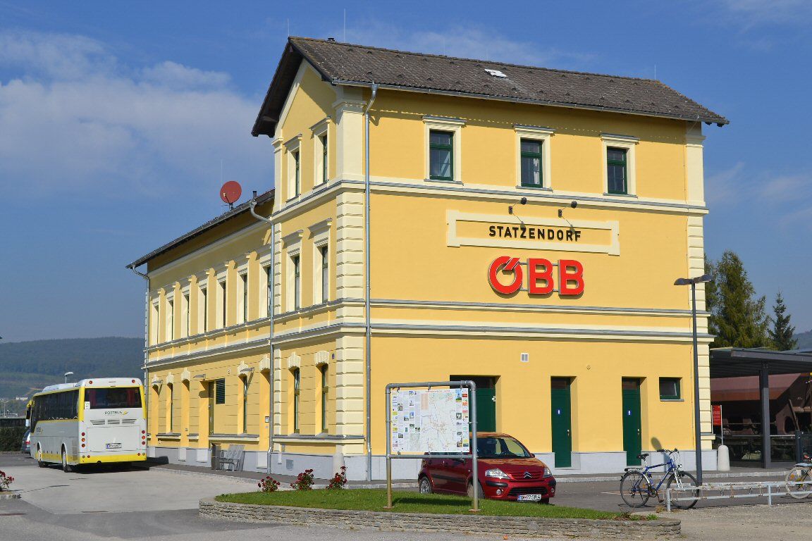 Sárga vasútállomás épület ÖBB logóval és autóbusszal az előtérben.
