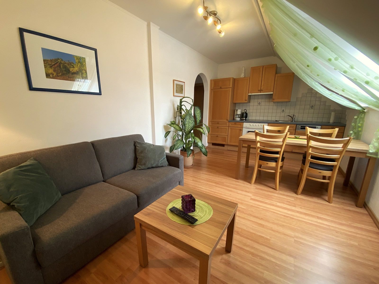 Nappali szürke kanapéval, fa asztallal, étkezőasztallal és konyhával a háttérben.