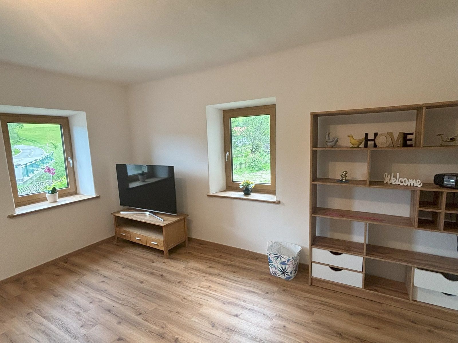 Egy nappali fapadlóval, egy asztalon lévő televízióval, két ablakkal és egy díszes polccal.