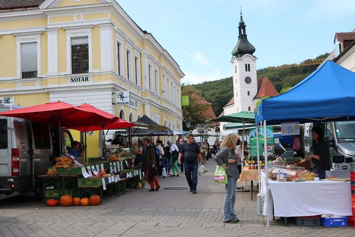 Élénk őstermelői piac bódékkal és látogatókkal egy sárga épület előtt, a háttérben egy templommal.