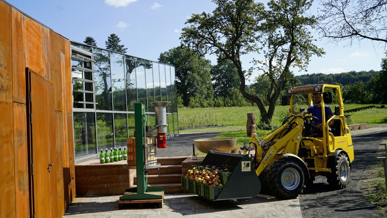 Egy sárga elektromos Hoftrac almás ládákat szállít egy modern épület előtt egy farmon.
