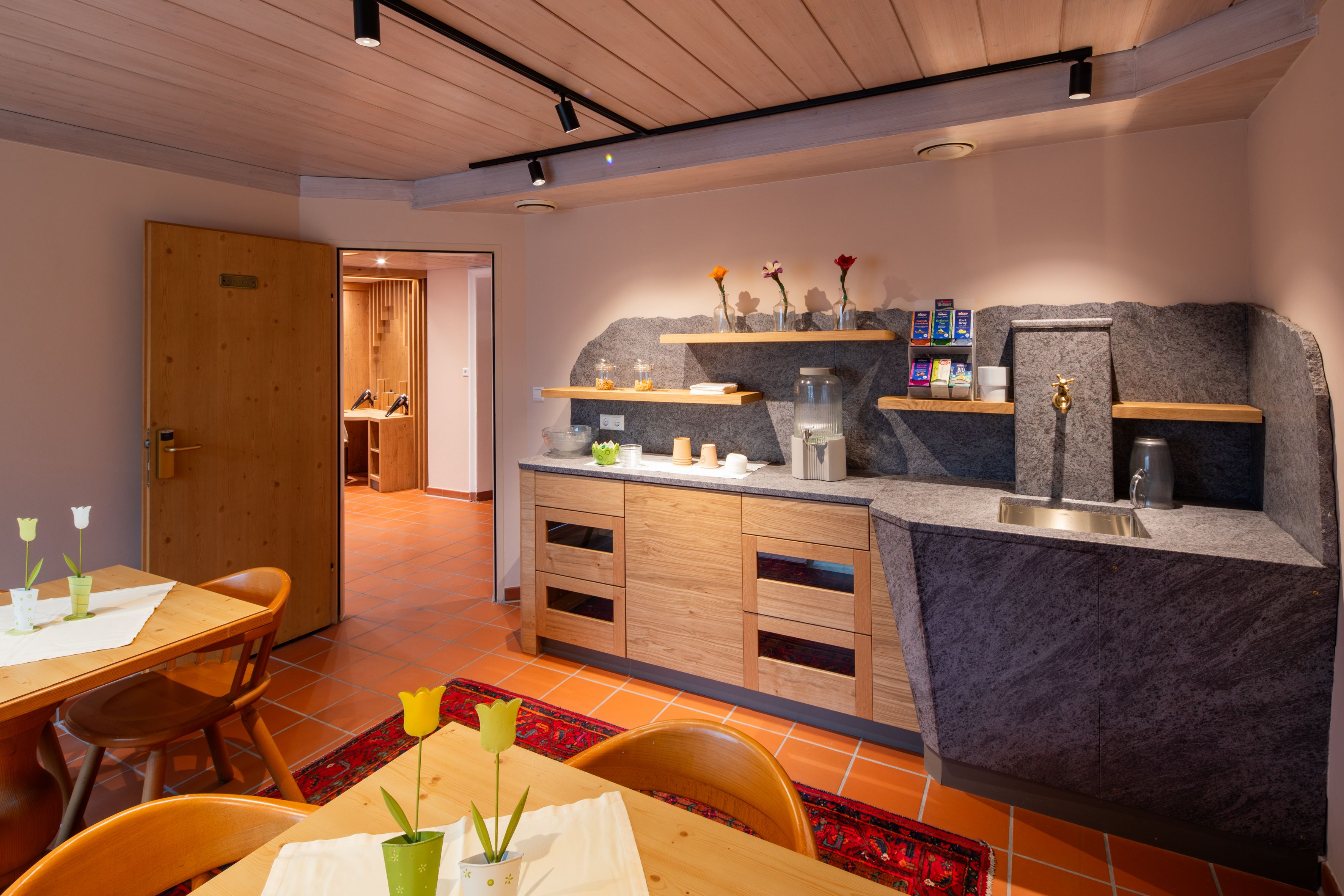 Hangulatos társalgó a szálloda fürdőjében, fabútorokkal, kis konyhasarokkal és dekoratív virágokkal.