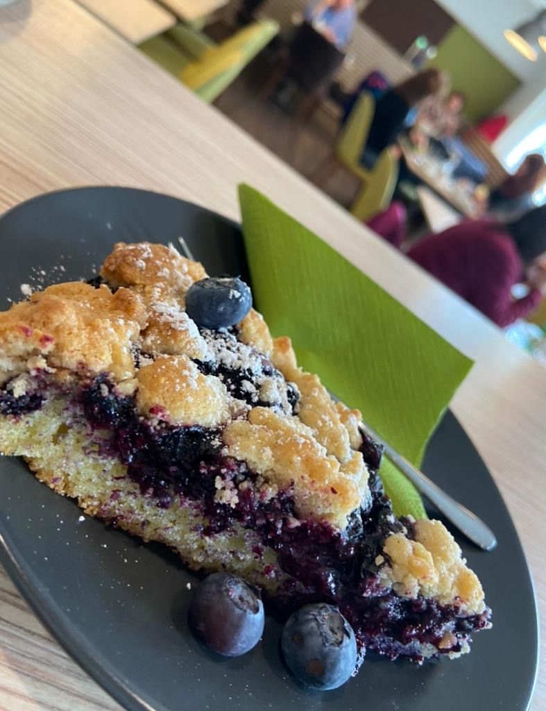Egy szelet áfonyás torta egy fekete tányéron, friss áfonyával és porcukorral, a háttérben egy elmosódott kávézójelenet.