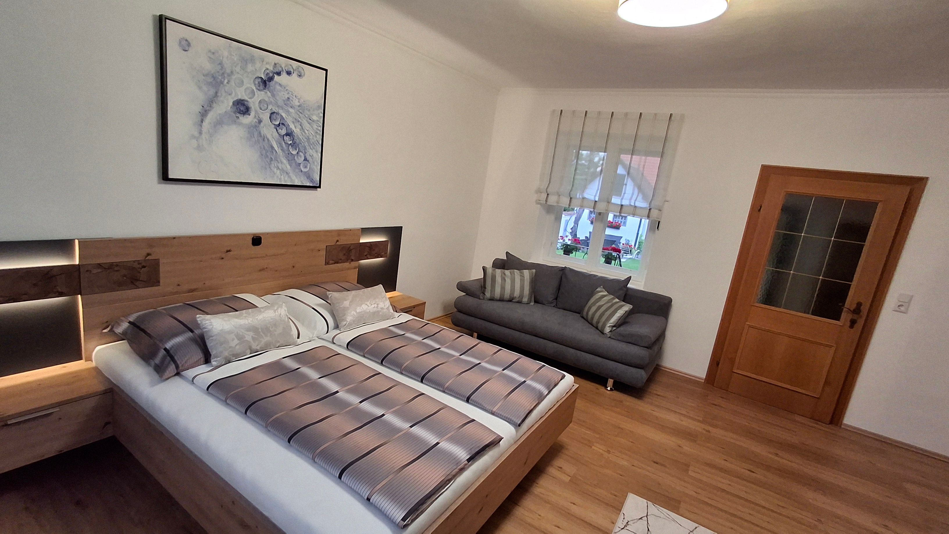 Modern hálószoba franciaággyal, szürke kanapéval, fapadlóval és absztrakt műalkotásokkal a falon.