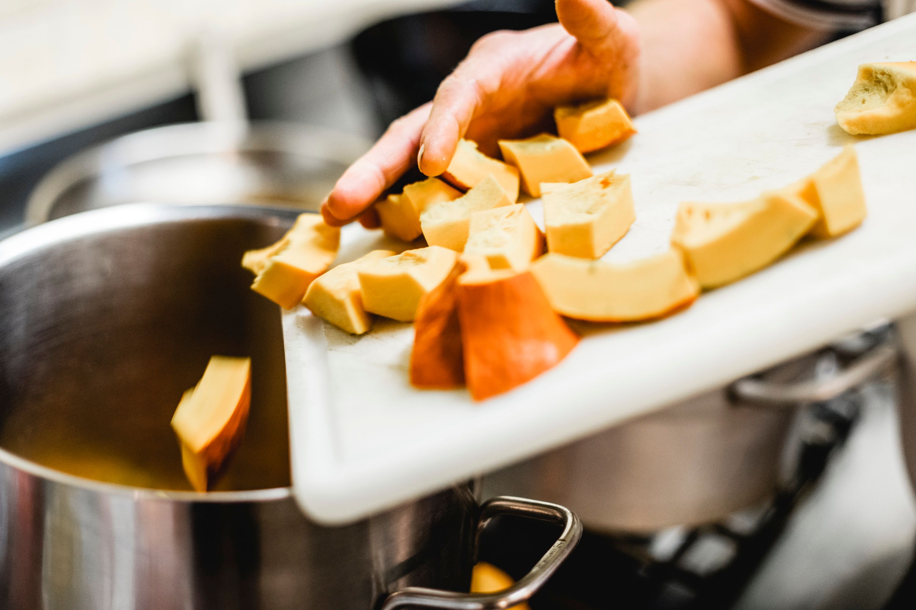 Egy kéz a vágódeszkáról egy fazékba tolja a szeletelt tökdarabokat.