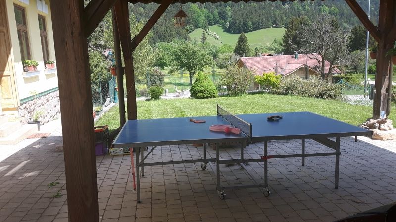 Asztalitenisz asztal egy kocsibeálló alatt, kilátással a zöld kertre és a háttérben lévő dombokra.