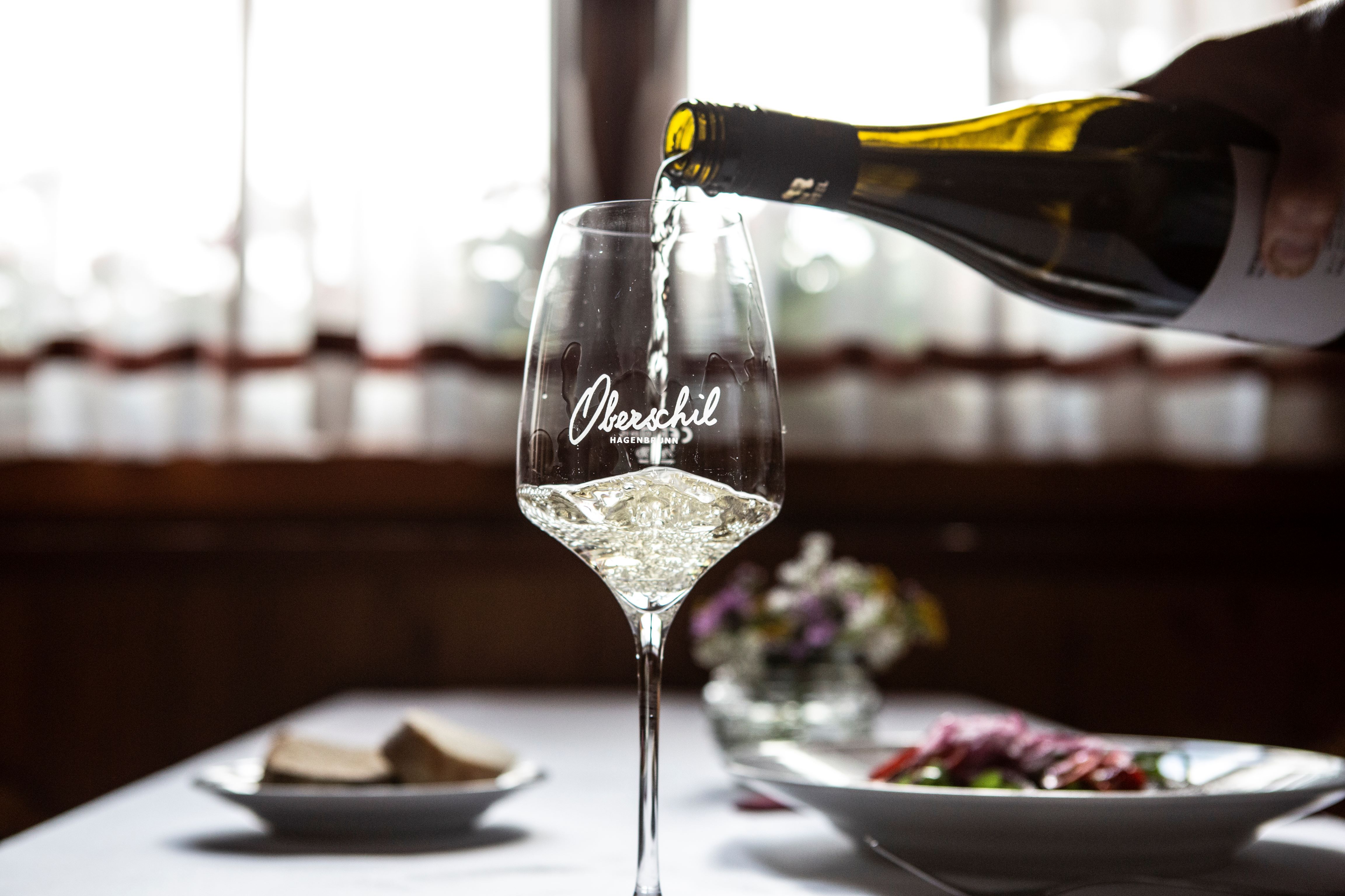 Egy pohár fehérbort töltenek, a háttérben virágokkal megterített asztal.