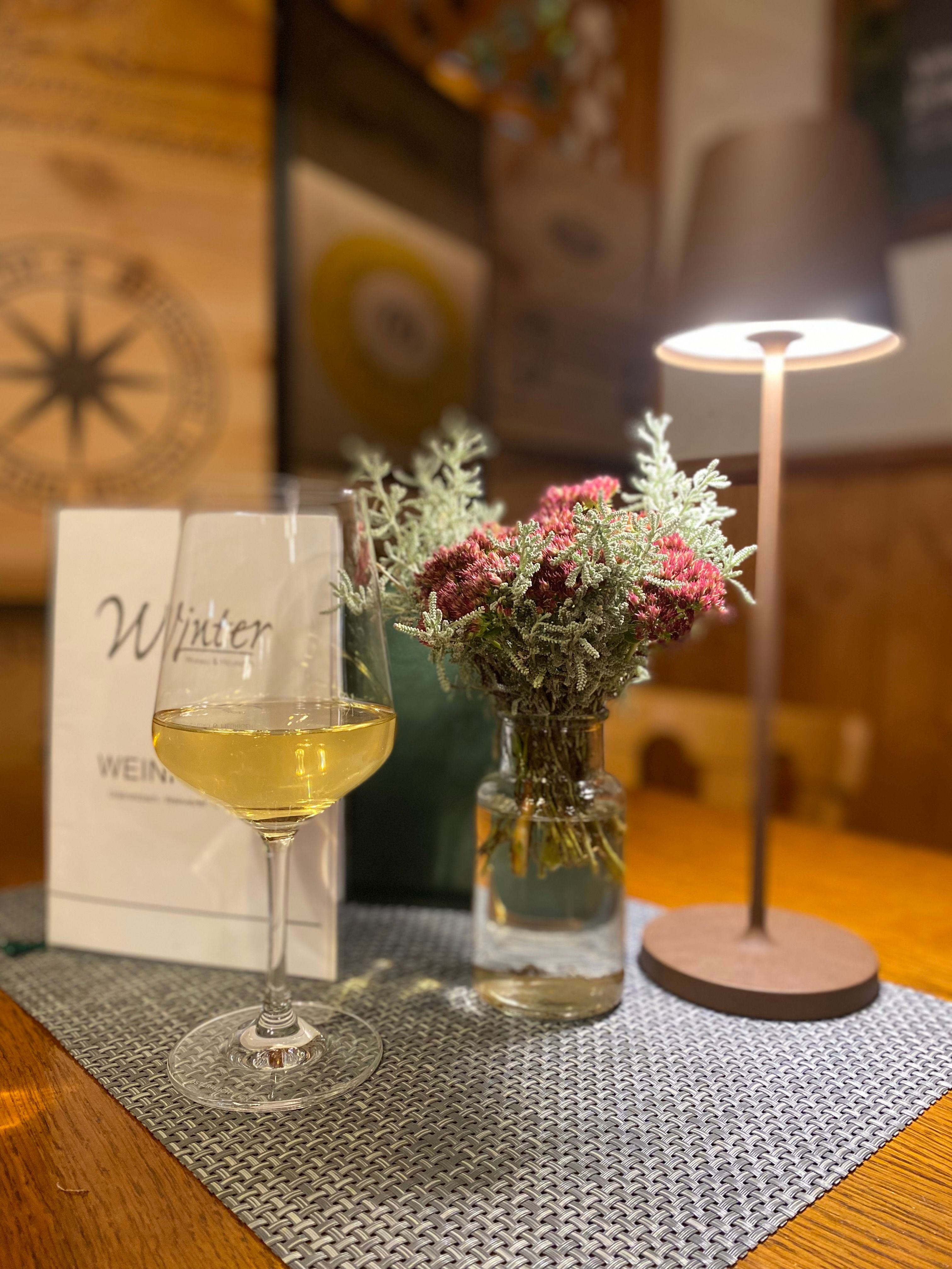 Egy fehérboros pohár áll az asztalon egy virágváza és egy asztali lámpa mellett.