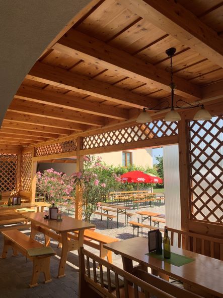 Fából készült pergola asztalokkal és padokkal, kilátás a kertre, piros napernyőkkel.