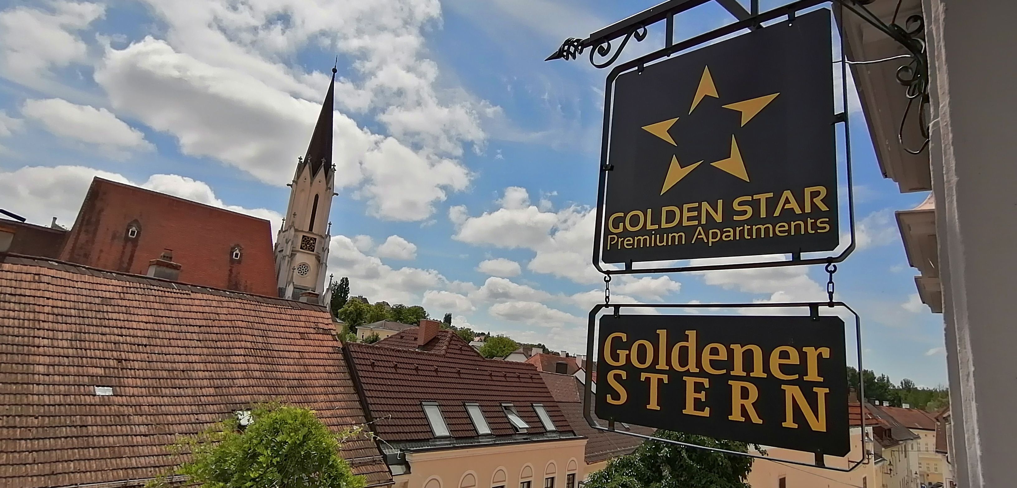 Kilátás egy "Golden Star Premium Apartments" és "Goldener Stern" feliratú táblára egy templomtorony és háztetők előtt.
