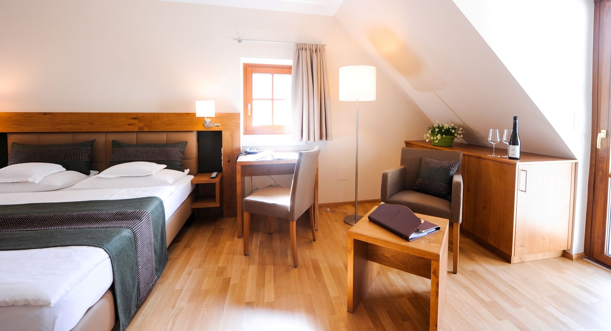 Hangulatos szállodai szoba franciaággyal, íróasztallal, fotelrel és egy üveg borral a szekrényen.