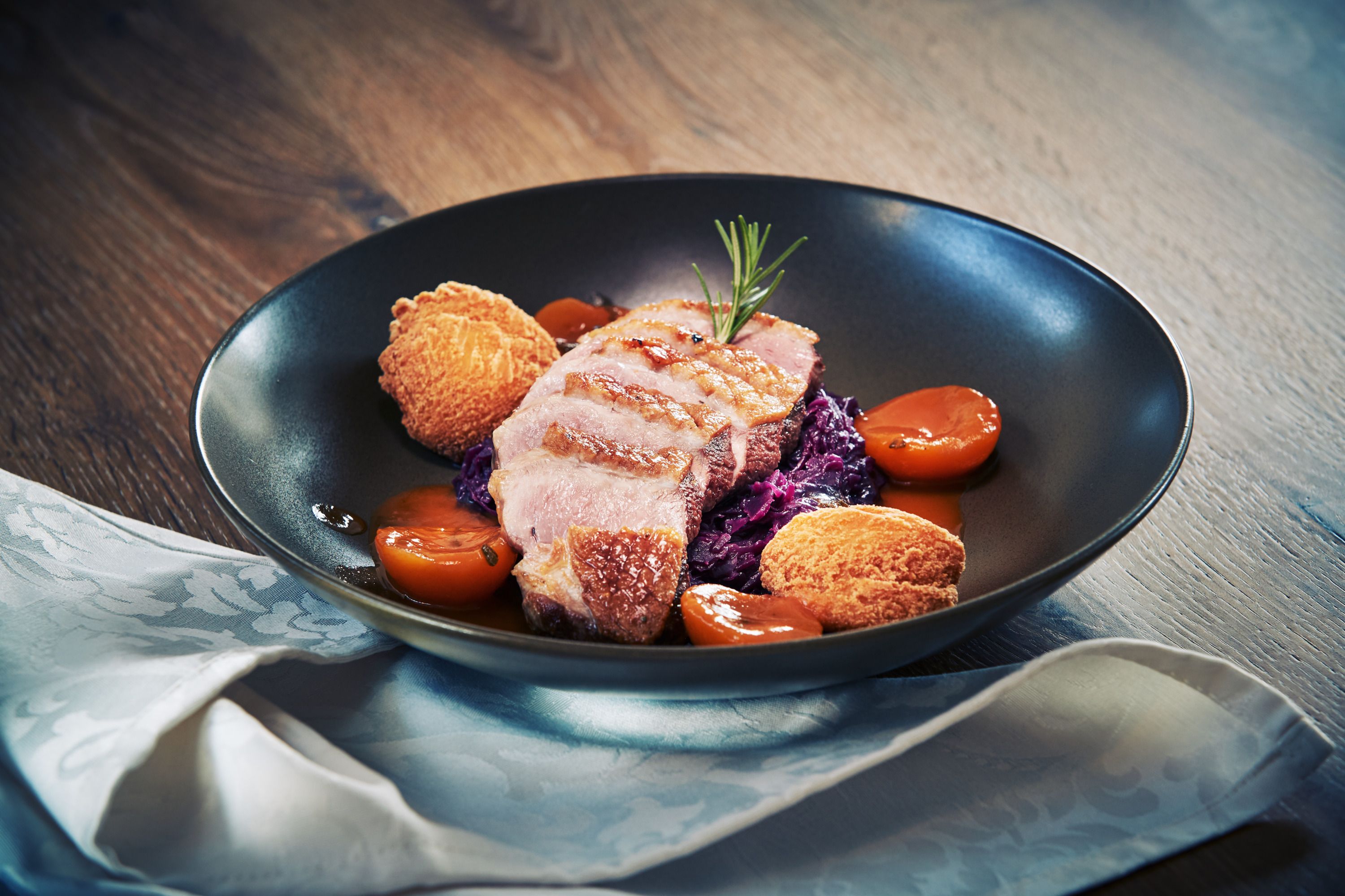 Gourmet étel kacsamell, vörös káposzta, burgonyagombóc és sárgabarack fekete tányéron.