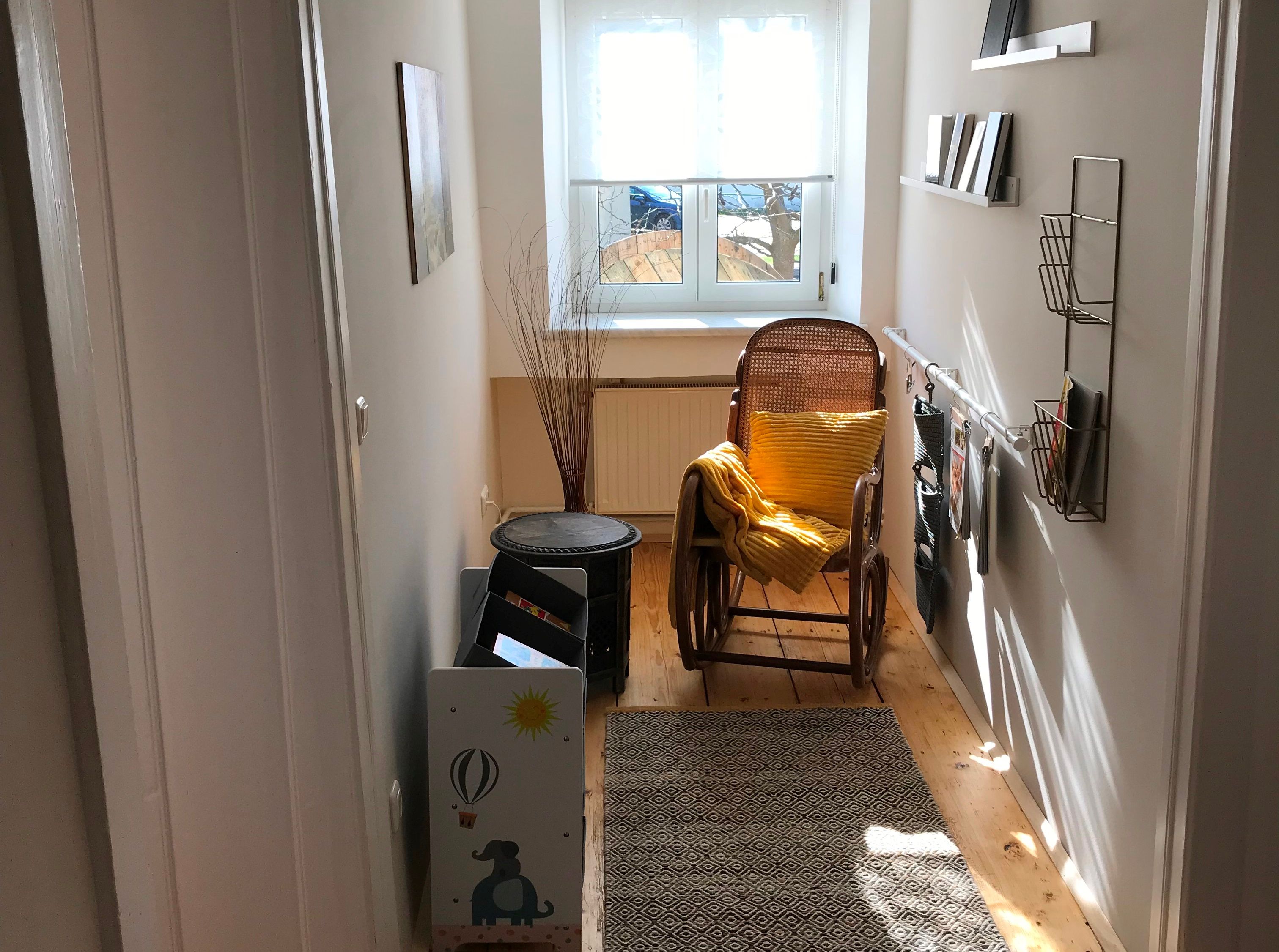 Egy keskeny bejárati rész hintaszékkel, sárga párnákkal, szőnyeggel és polcokkal a fal mellett. Egy ablak a folyosó végén.