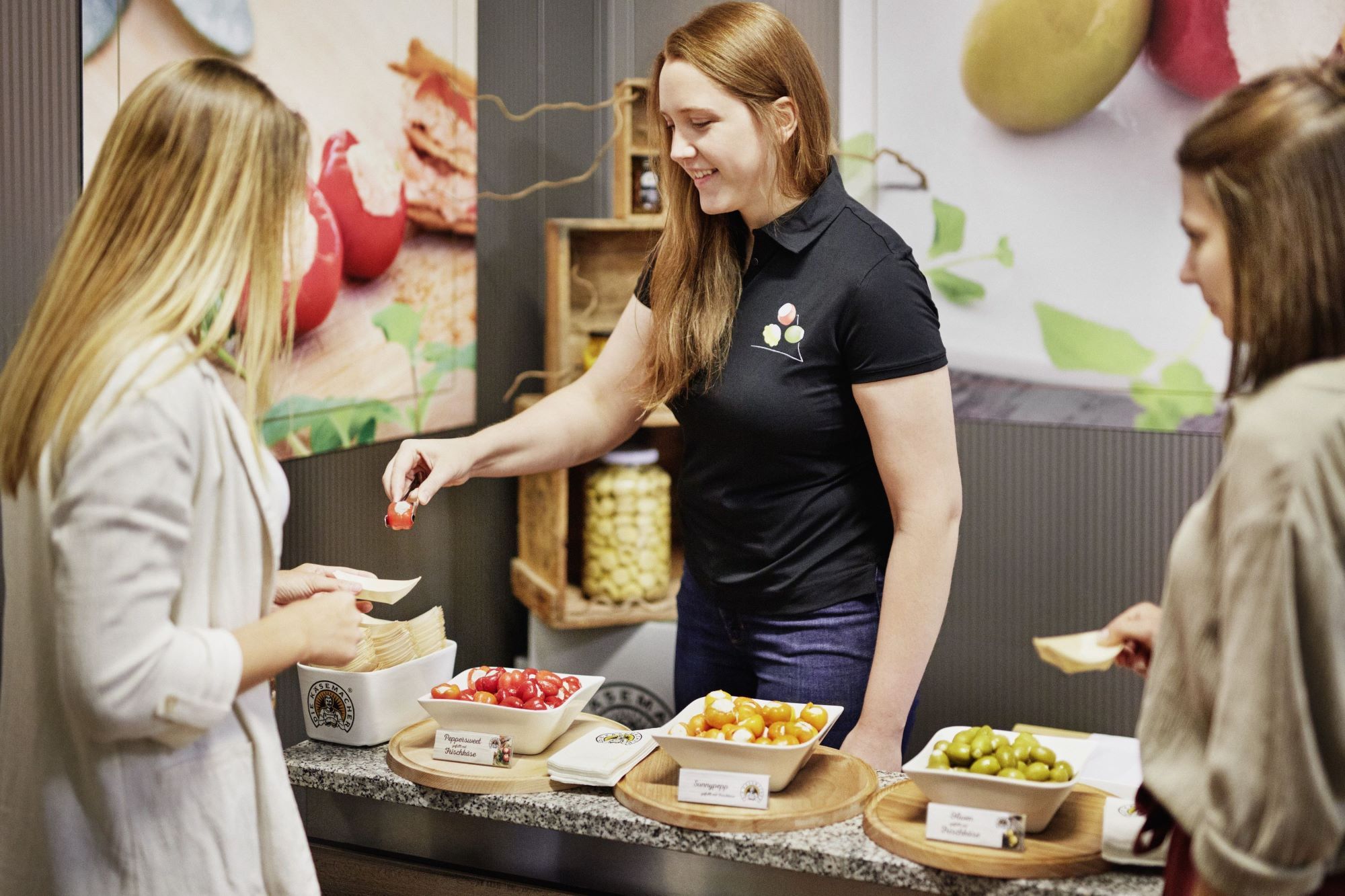 Egy nő két másik nőnek különböző töltött paprikákat és olajbogyót kínál kóstolásra.