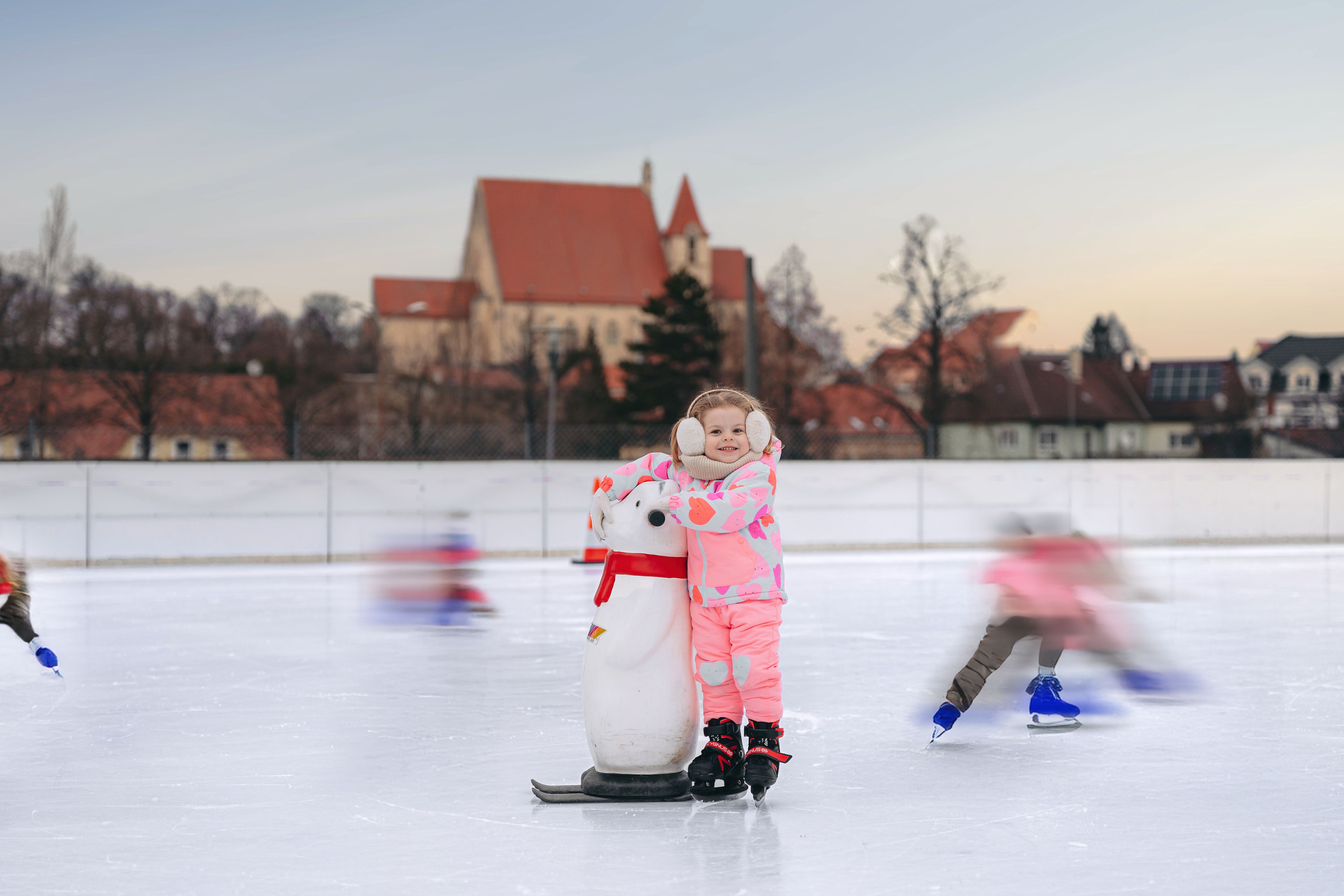 Egy rózsaszín ruhás gyermek áll a jégpályán egy pingvin segédeszközzel. A háttérben egy templom látható.