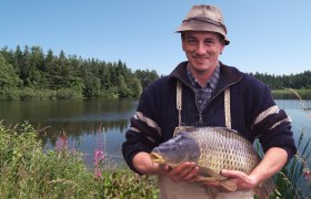 Egy f&eacute;rfi egy nagy pontyot tart egy t&oacute; előtt, a h&aacute;tt&eacute;rben egy erdővel.
