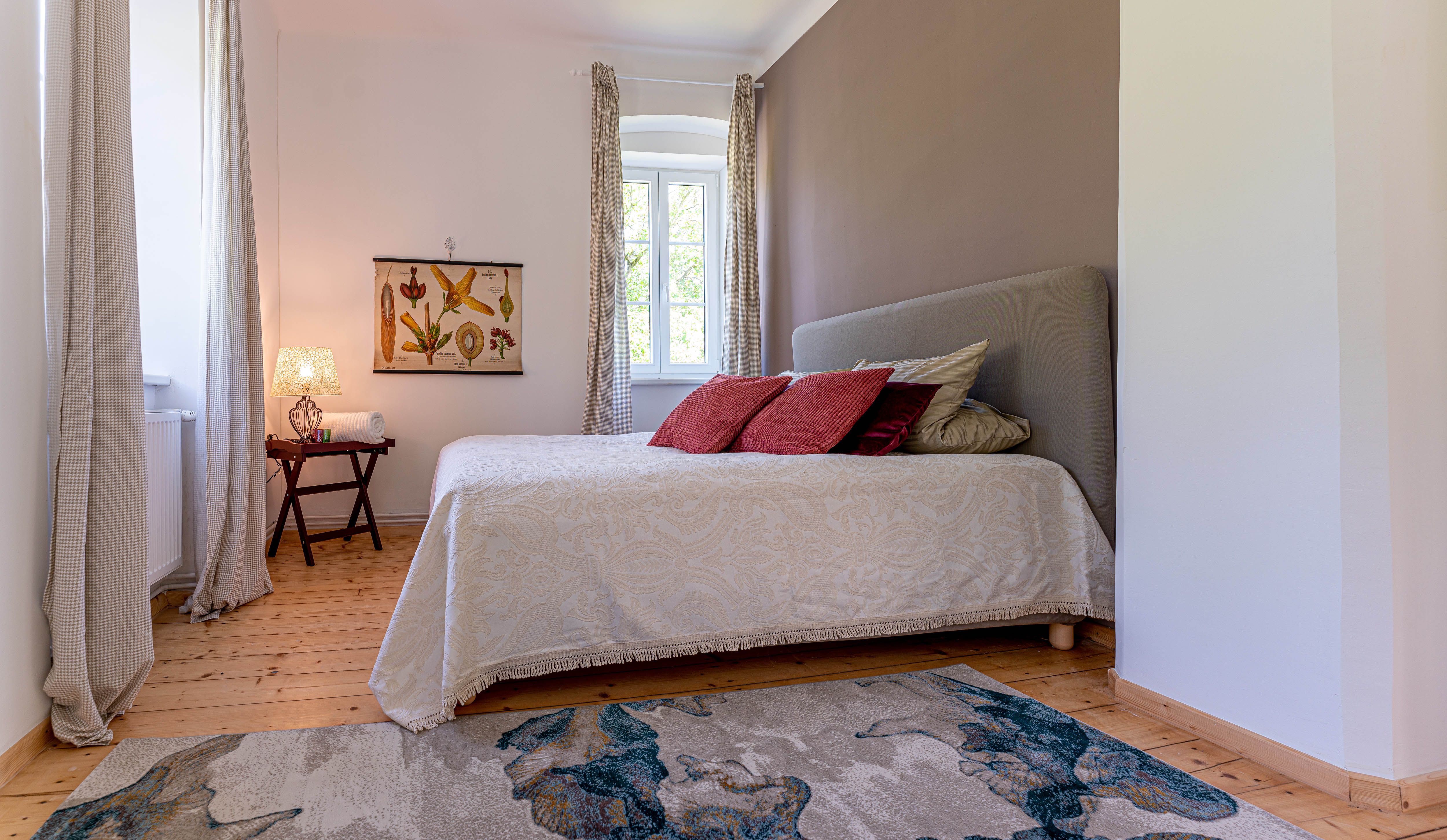 Hálószoba ággyal, piros párnákkal, szőnyeggel és függönyös ablakkal.