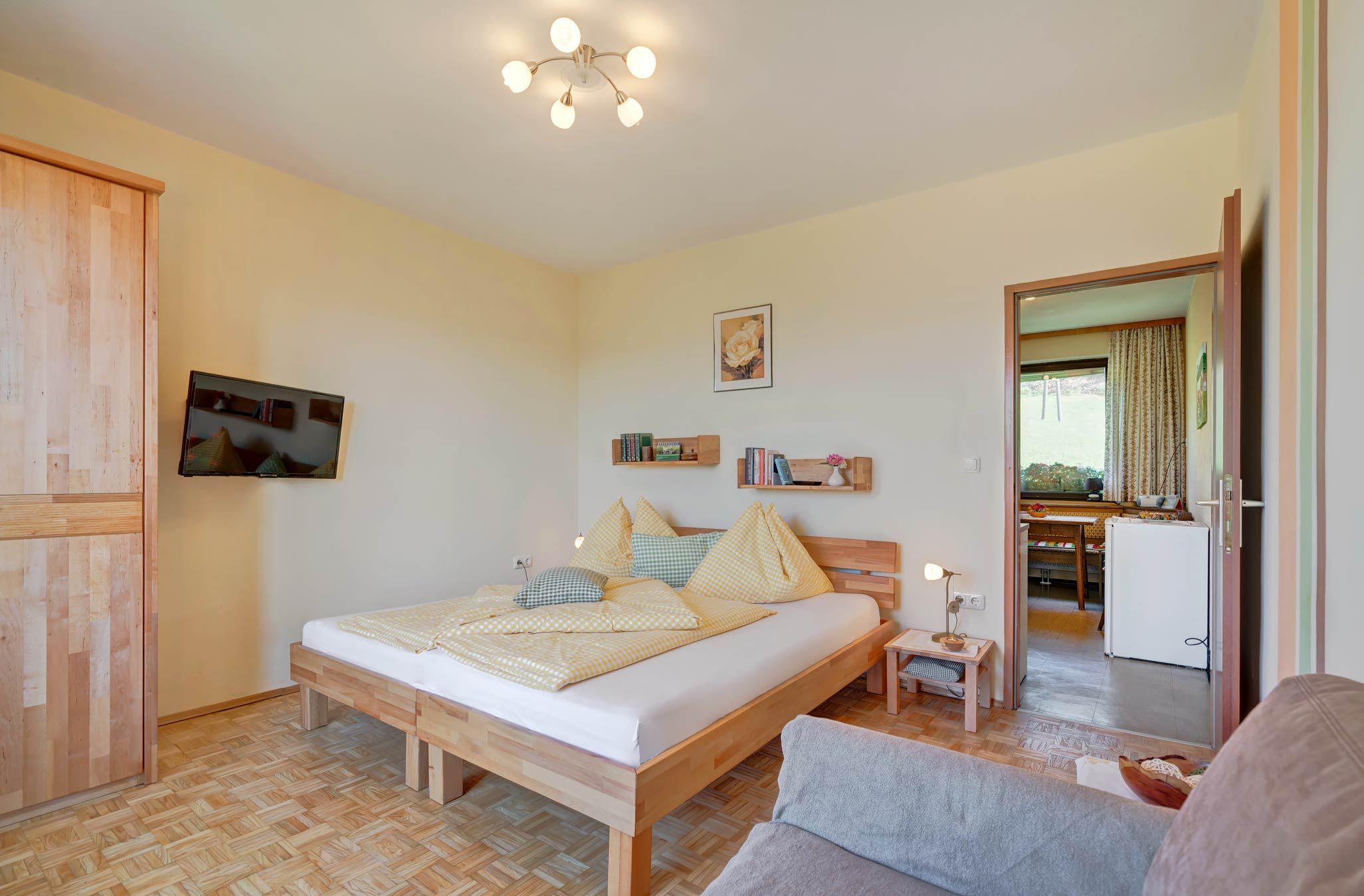 Hangulatos hálószoba fapadlóval, franciaággyal, sárga ágyneművel, síkképernyős TV-vel a falon és nyitott átjáróval a konyhába.