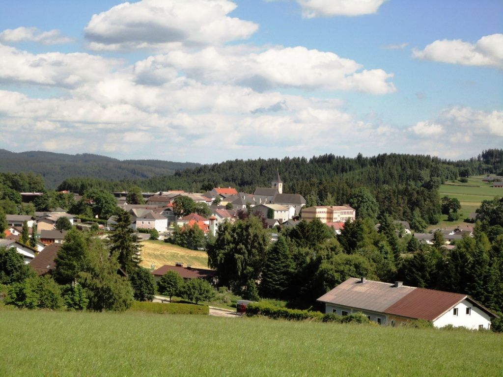 Panoráma Martinsberg mezővárosáról, a templomról és a környező házakról, erdőkkel és mezőkkel körülvéve, kék ég alatt.