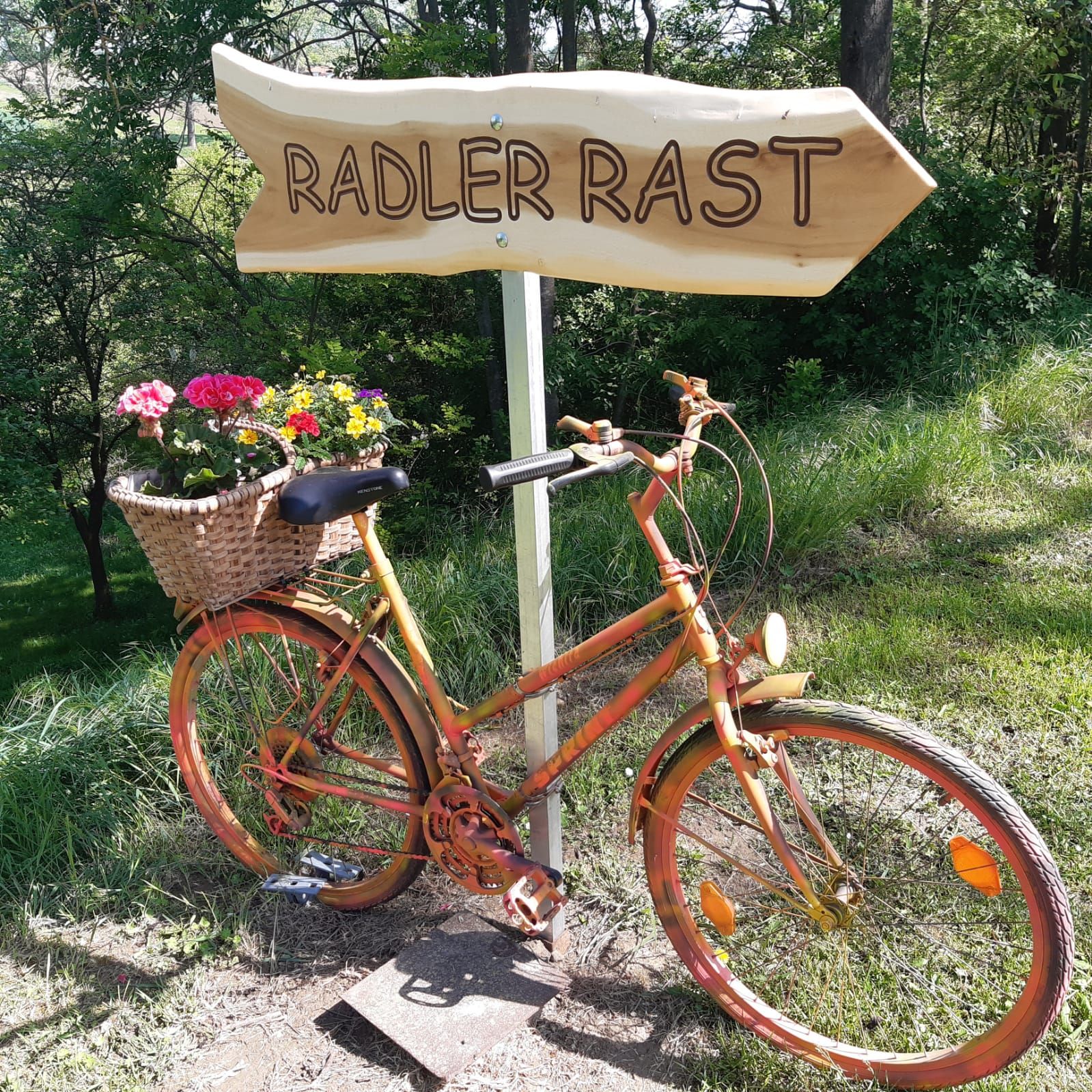 Egy narancssárga kerékpár, rajta egy virágkosárral, egy „Radler Rast” feliratú fa táblánál áll zöld környezetben.
