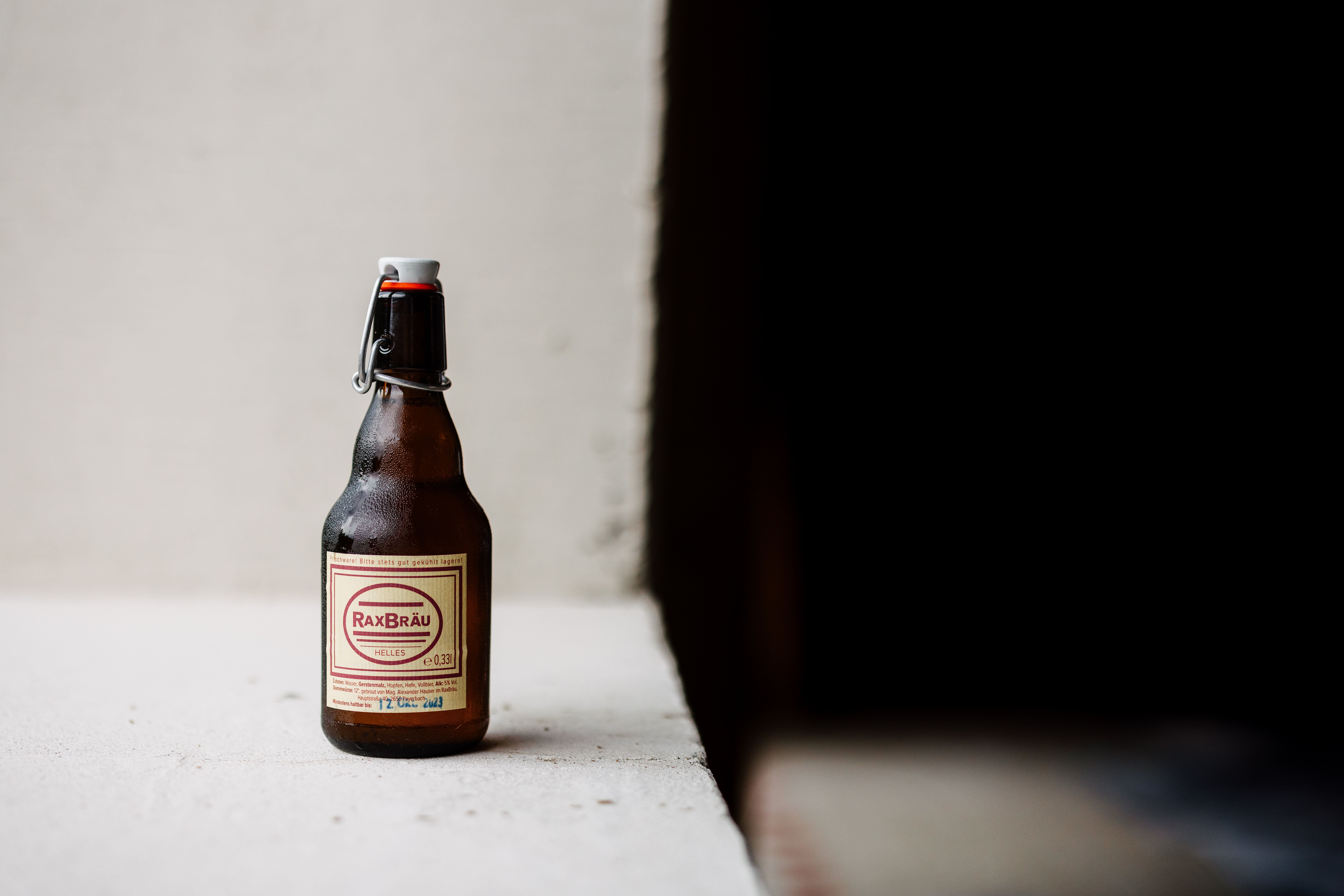 Egy üveg RaxBräu Helles Bier fehér felületen, sötét háttér előtt.
