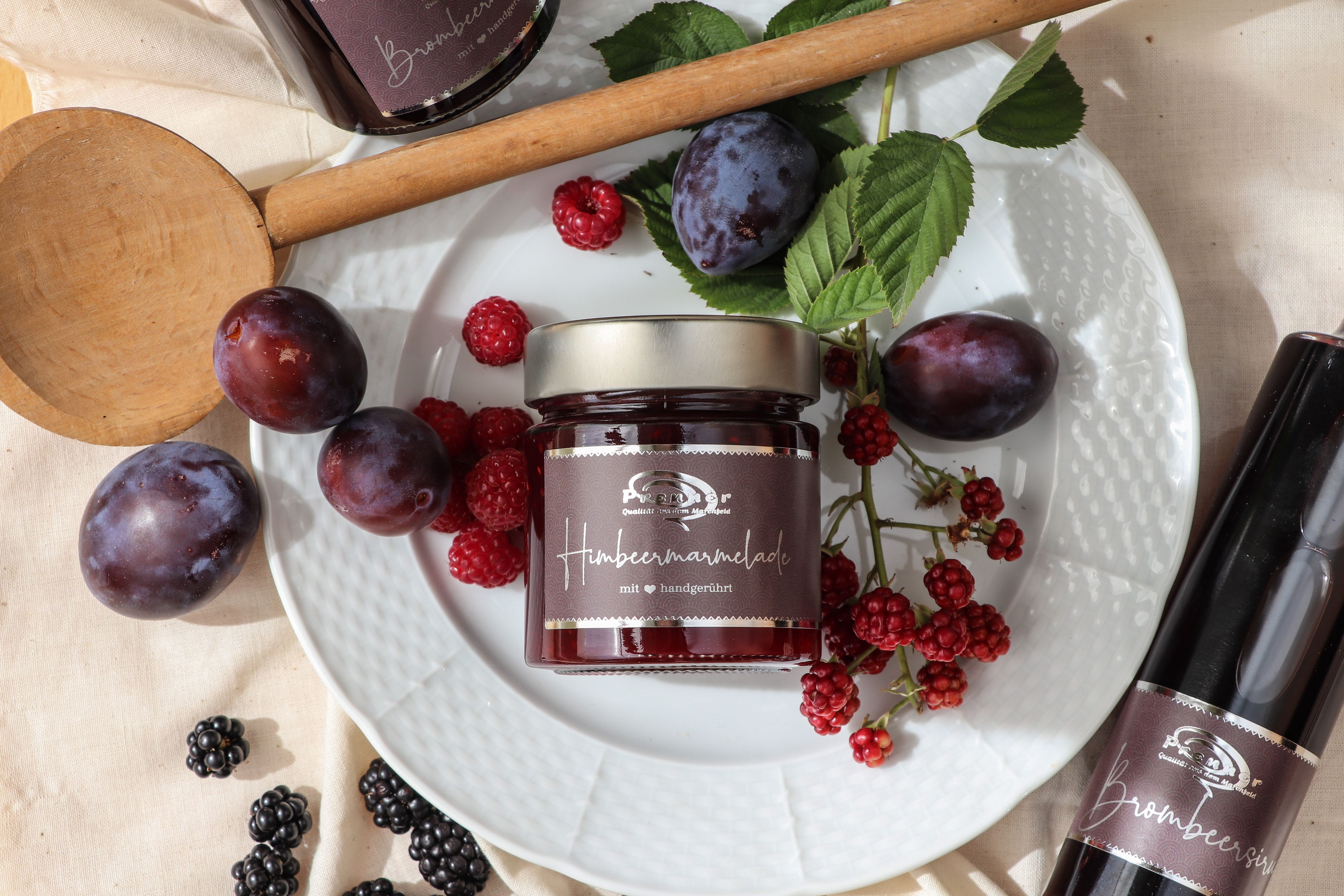 Egy üveg málnalekvár egy tányéron friss szilvával, málnával és szederrel, körülötte egy fakanál és egy üveg szeder szirup.