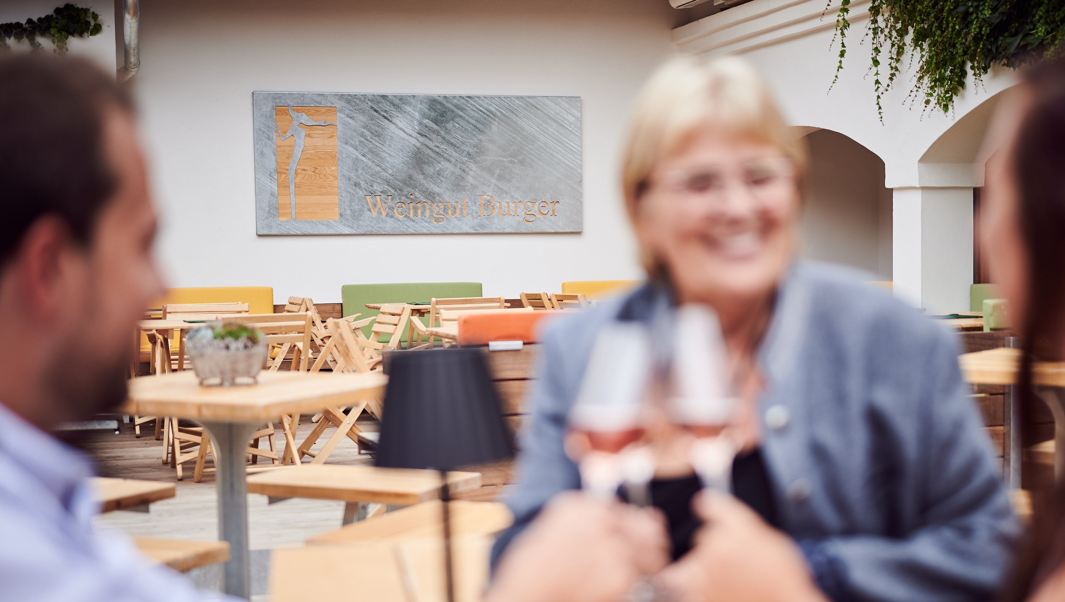 Három ember koccint egy pohár borral egy udvaron, a háttérben egy "Weingut Burger" feliratú táblával.