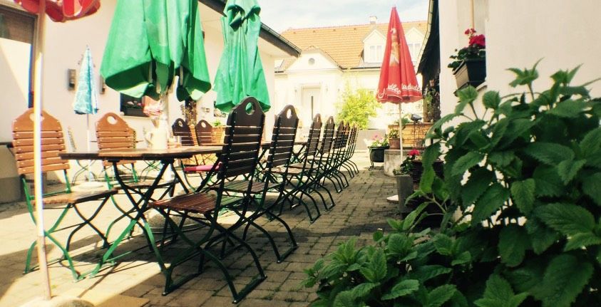 Egy üres vendégkert faasztalokkal és székekkel, zöld napernyőkkel és növényekkel körülvéve.