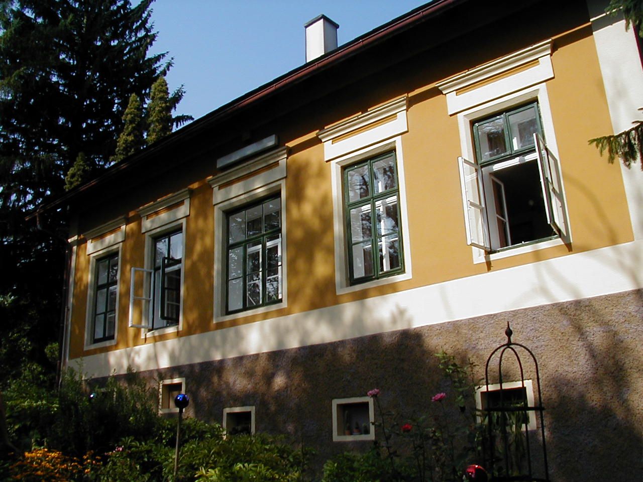 Sárga ház nyitott ablakokkal és kerttel az előtérben.