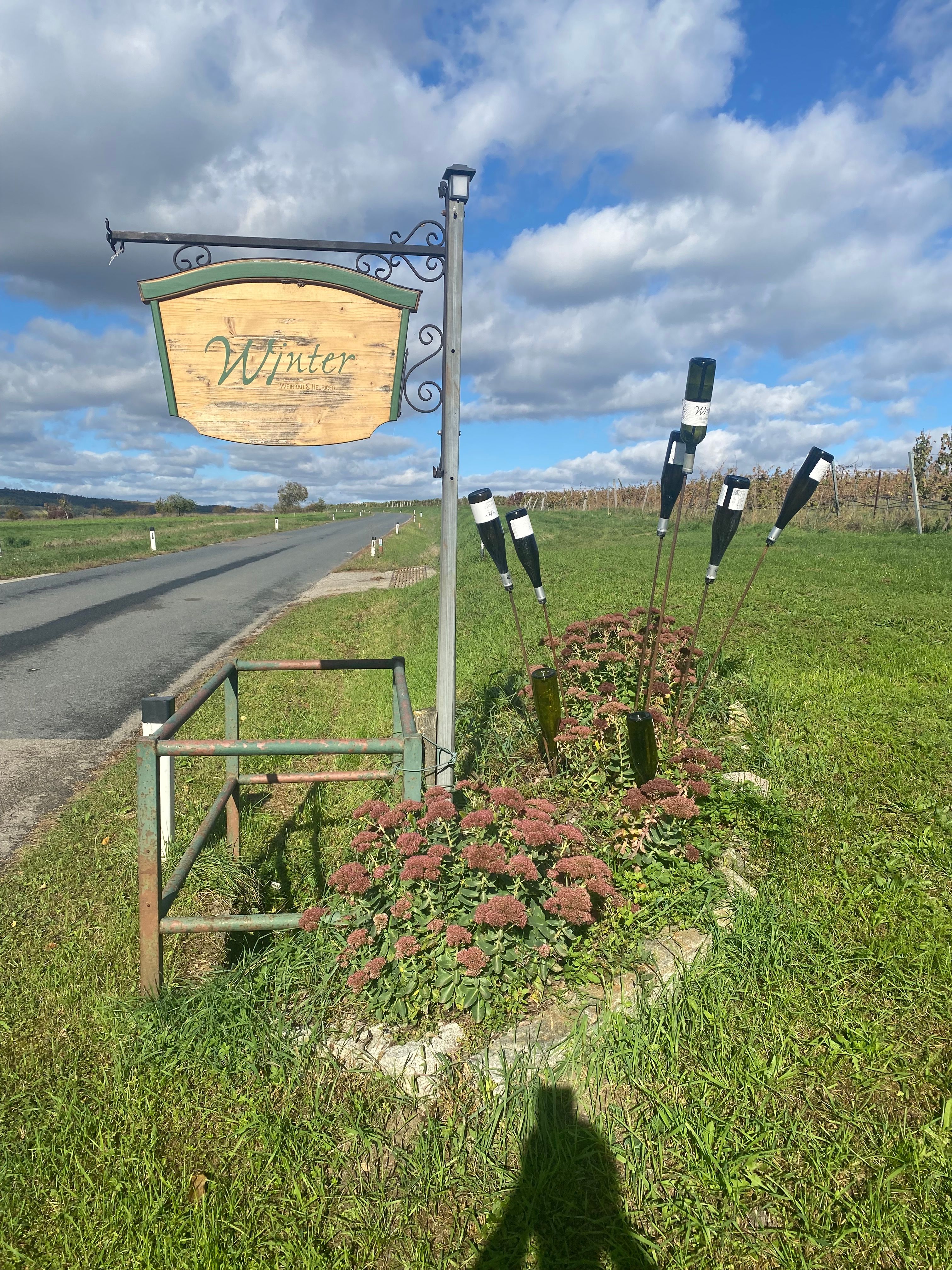 Az út szélén egy fából készült "Tél" feliratú tábla áll egy rét mellett, virágokkal és palackokkal díszítve.