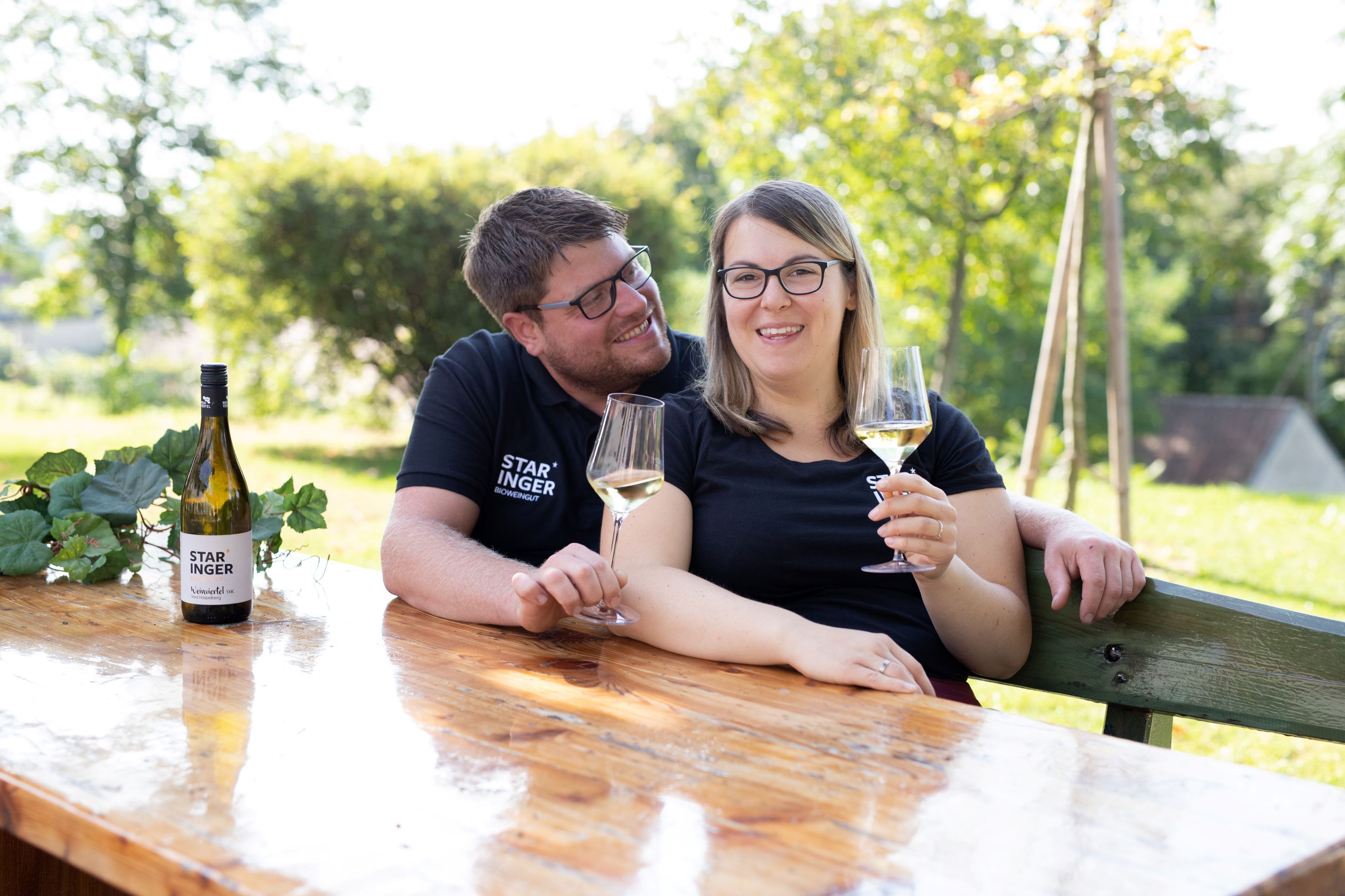 Egy házaspár ül egy faasztalnál odakint, kezükben pohár borral. Az asztalon egy üveg "Staringer" feliratú bor áll.