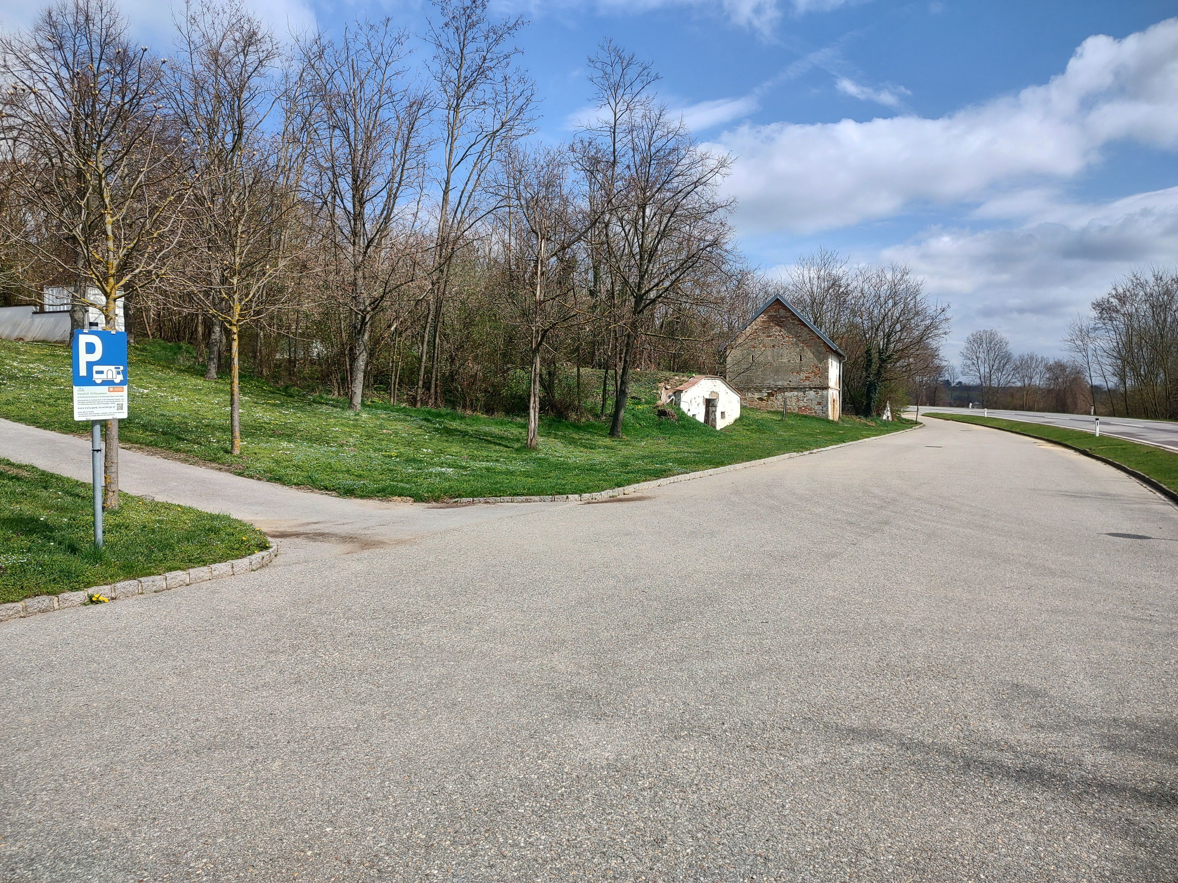 Autóparkoló lakóautó parkoló táblával, a háttérben fák és régi épület.