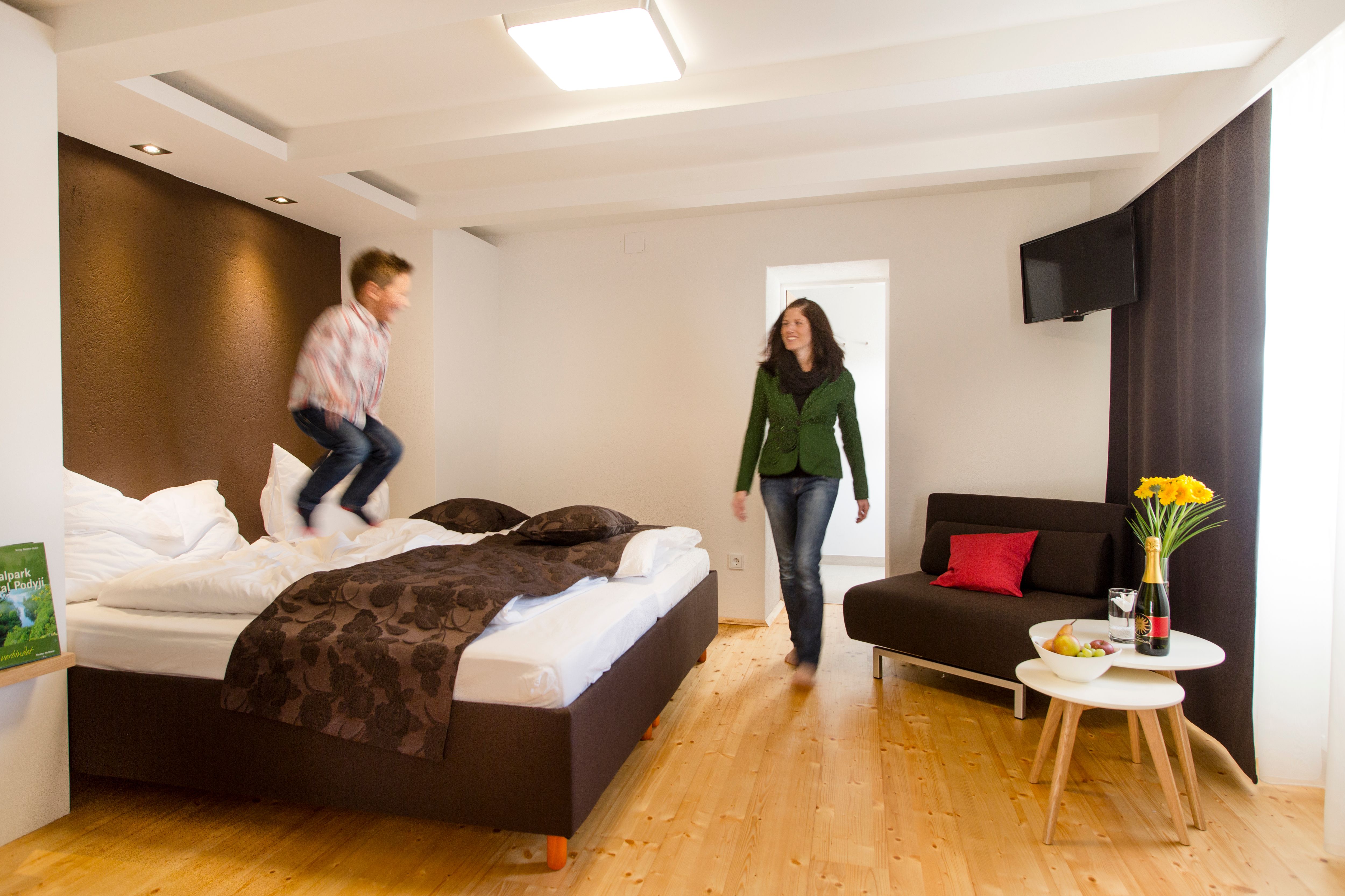 Egy modern hotelszobában egy gyermek ugrál az ágyon, miközben egy nő mosolyogva nézi. Mellette egy asztal virágokkal és borral áll.