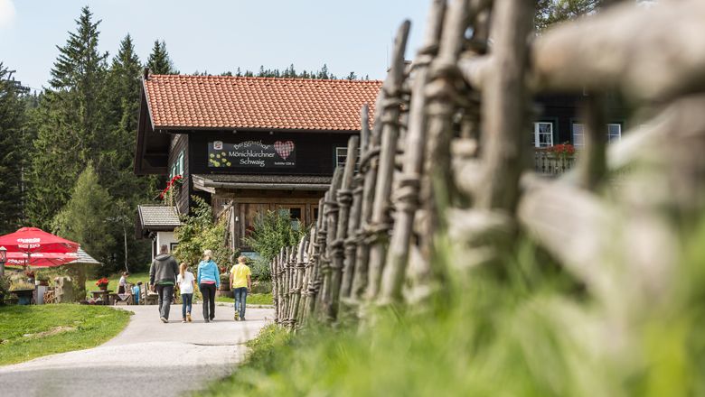 Három ember sétál egy hegyi fogadó felé, amelyet fakerítés és fák vesznek körül.
