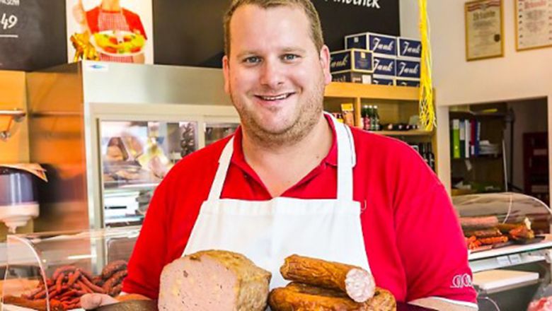 Egy hentes piros ingben és fehér kötényben különböző kolbásztermékeket tart egy hentesüzletben.