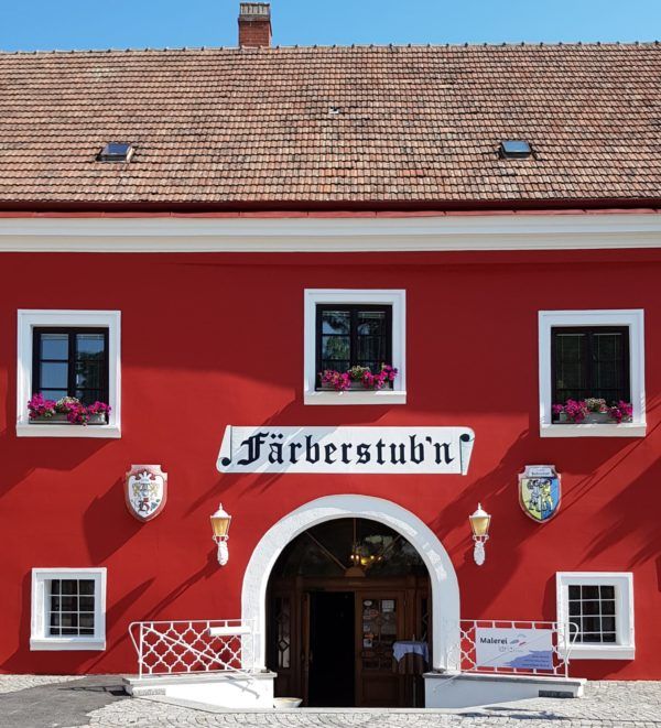 Piros épület a "Färberstub'n" felirattal és virágzó virágokkal az ablakokban.