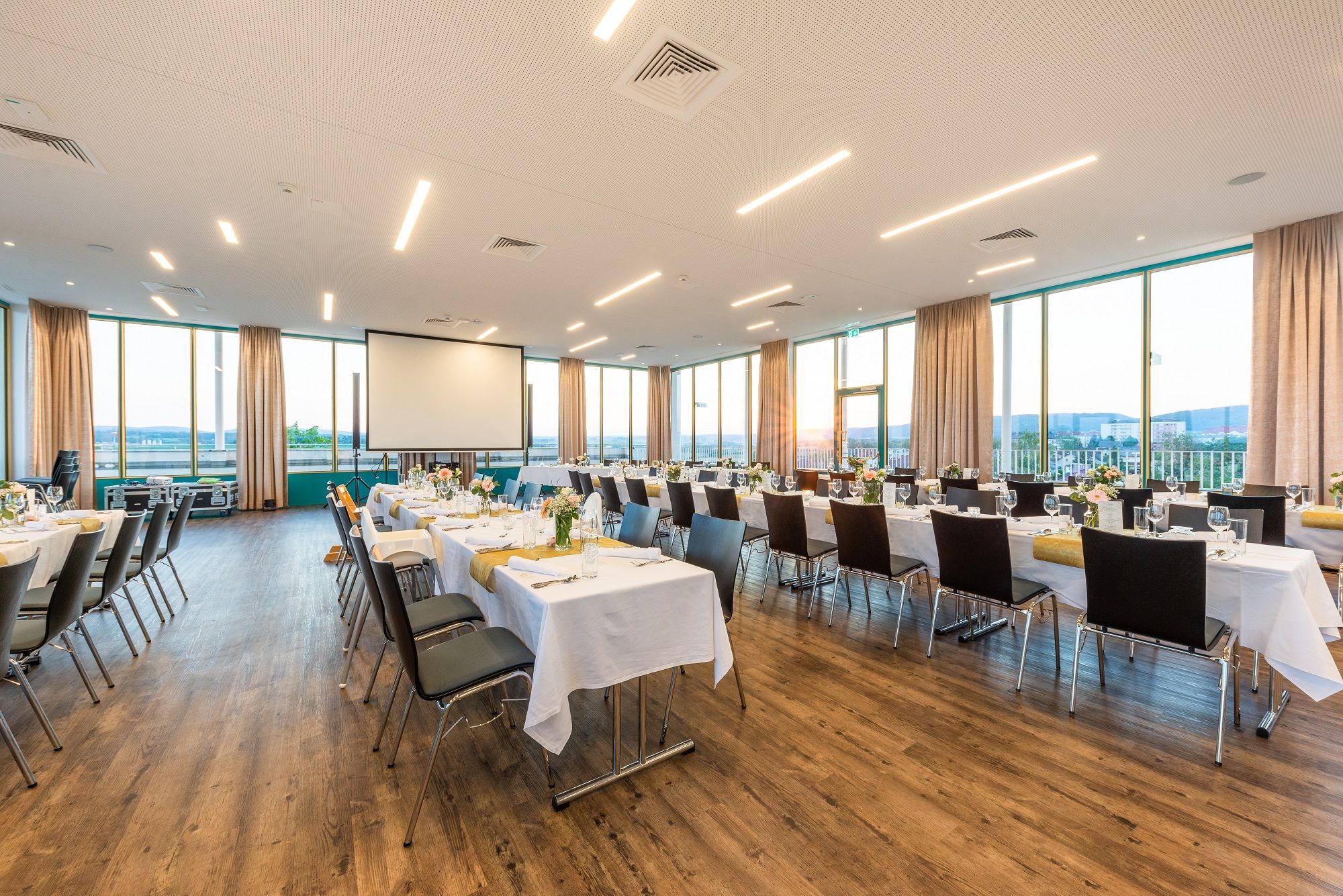 Nagy, világos terem, fedett asztalokkal és székekkel, nagy ablakokkal és kivetítővel.