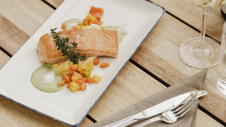 Egy t&aacute;ny&eacute;r s&uuml;lt lazac z&ouml;lds&eacute;gekkel &eacute;s z&ouml;ld m&aacute;rt&aacute;ssal, fűszern&ouml;v&eacute;nyekkel d&iacute;sz&iacute;tve, egy faasztalon, evőeszk&ouml;z&ouml;kkel &eacute;s borospoh&aacute;rral.