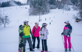 S&iacute;elők &eacute;s snowboardosok csoportja egy havas p&aacute;ly&aacute;n, a h&aacute;tt&eacute;rben erdő.