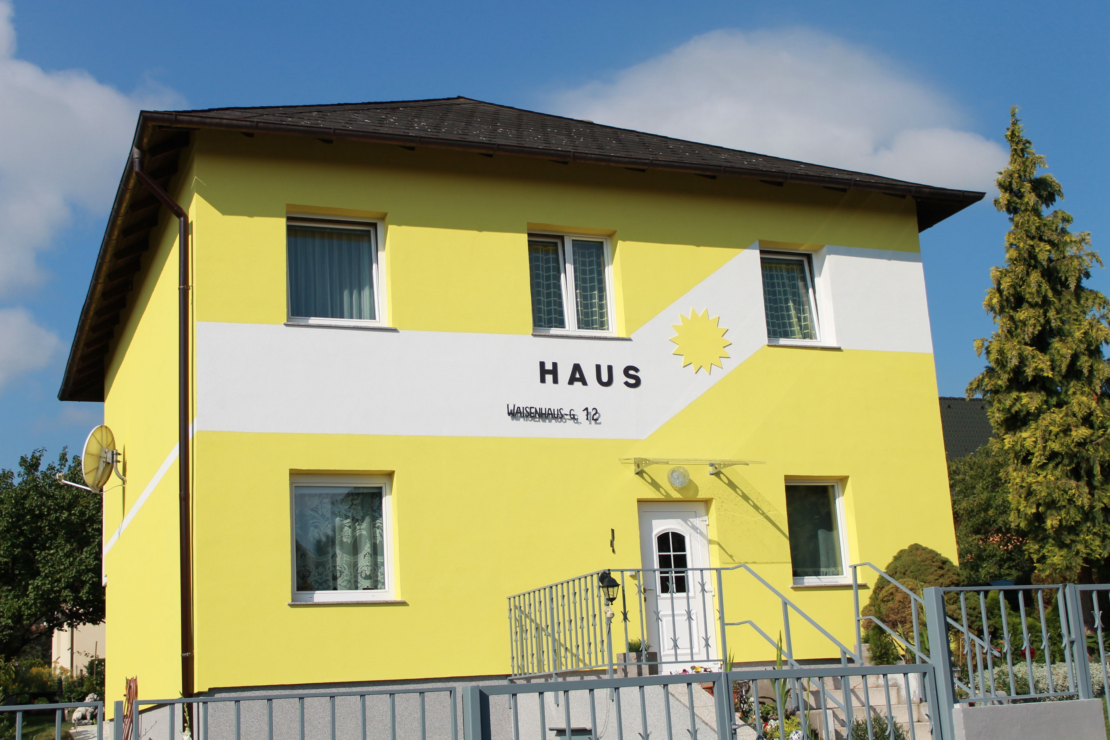 Sárga ház, homlokzatán a "Haus" felirat és egy nap.
