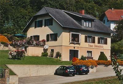 A "Haus Donaublick" feliratú kétszintes ház, amelyet virágok és fák vesznek körül. A ház előtt két autó parkol.