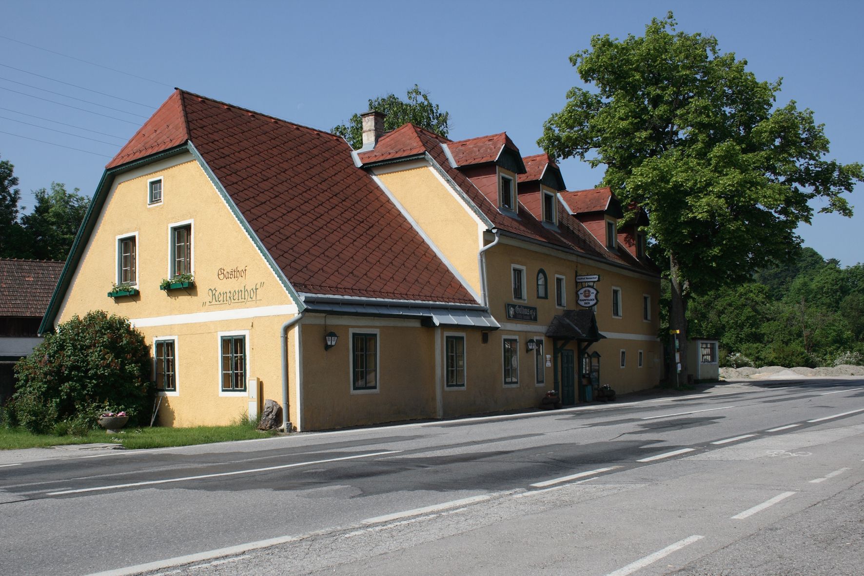 Sárga, piros tetejű fogadó egy út mentén, fákkal körülvéve.