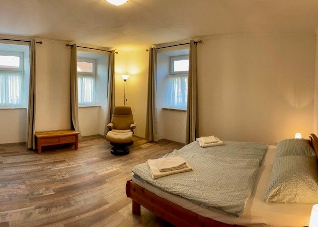 Egyszerű hálószoba franciaággyal, fotel, éjjeliszekrény és három ablak függönnyel.