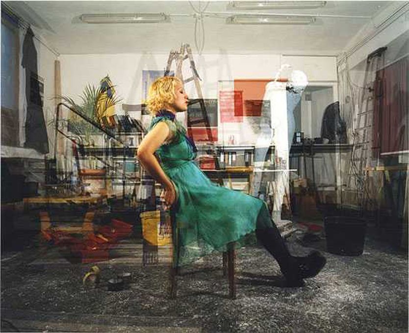 Egy zöld ruhás nő ül egy széken egy zsúfolt szobában.