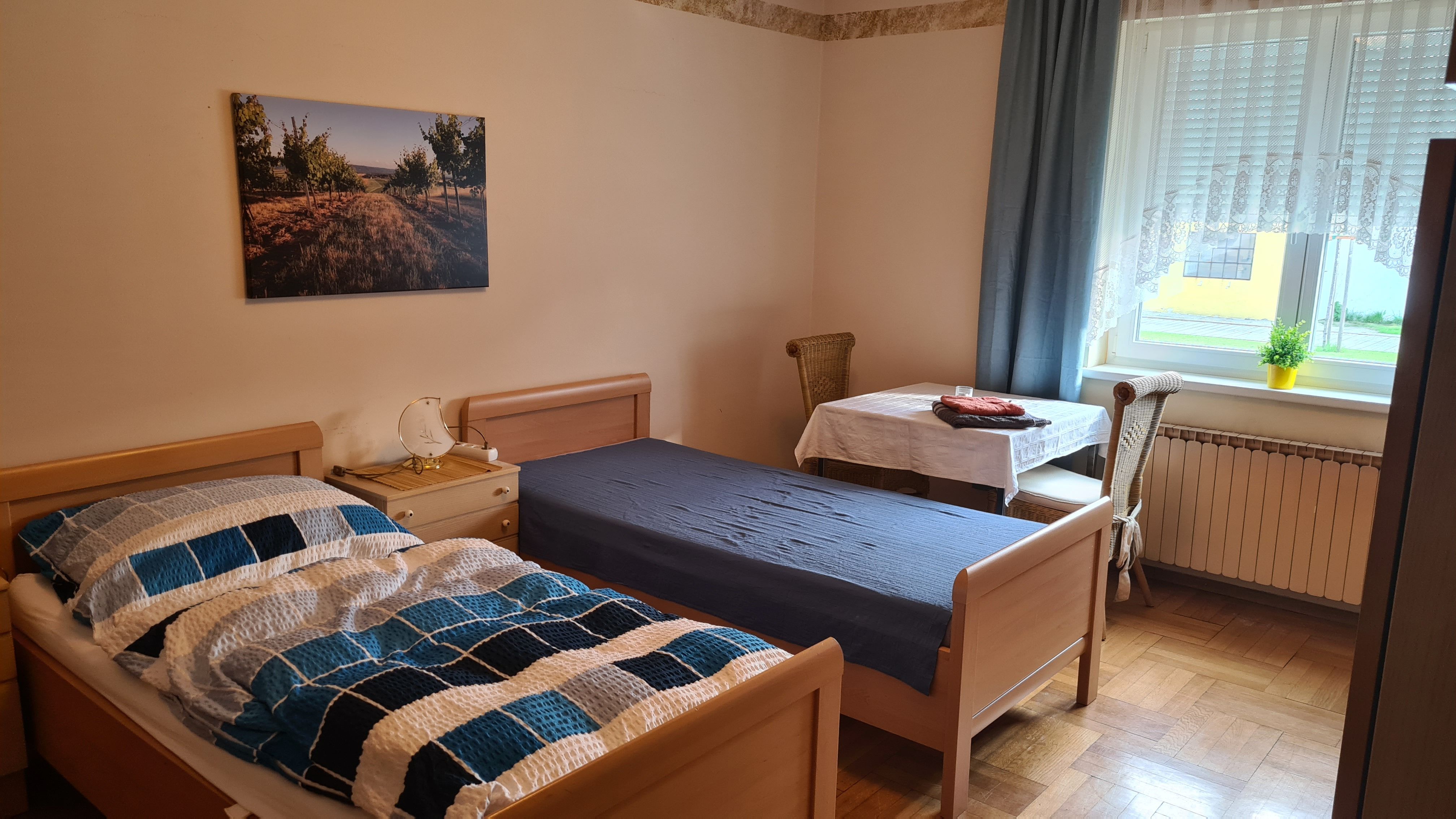 Egyszerűen berendezett hálószoba két egyszemélyes ággyal, asztallal és székkel, valamint egy függönyös ablakkal.
