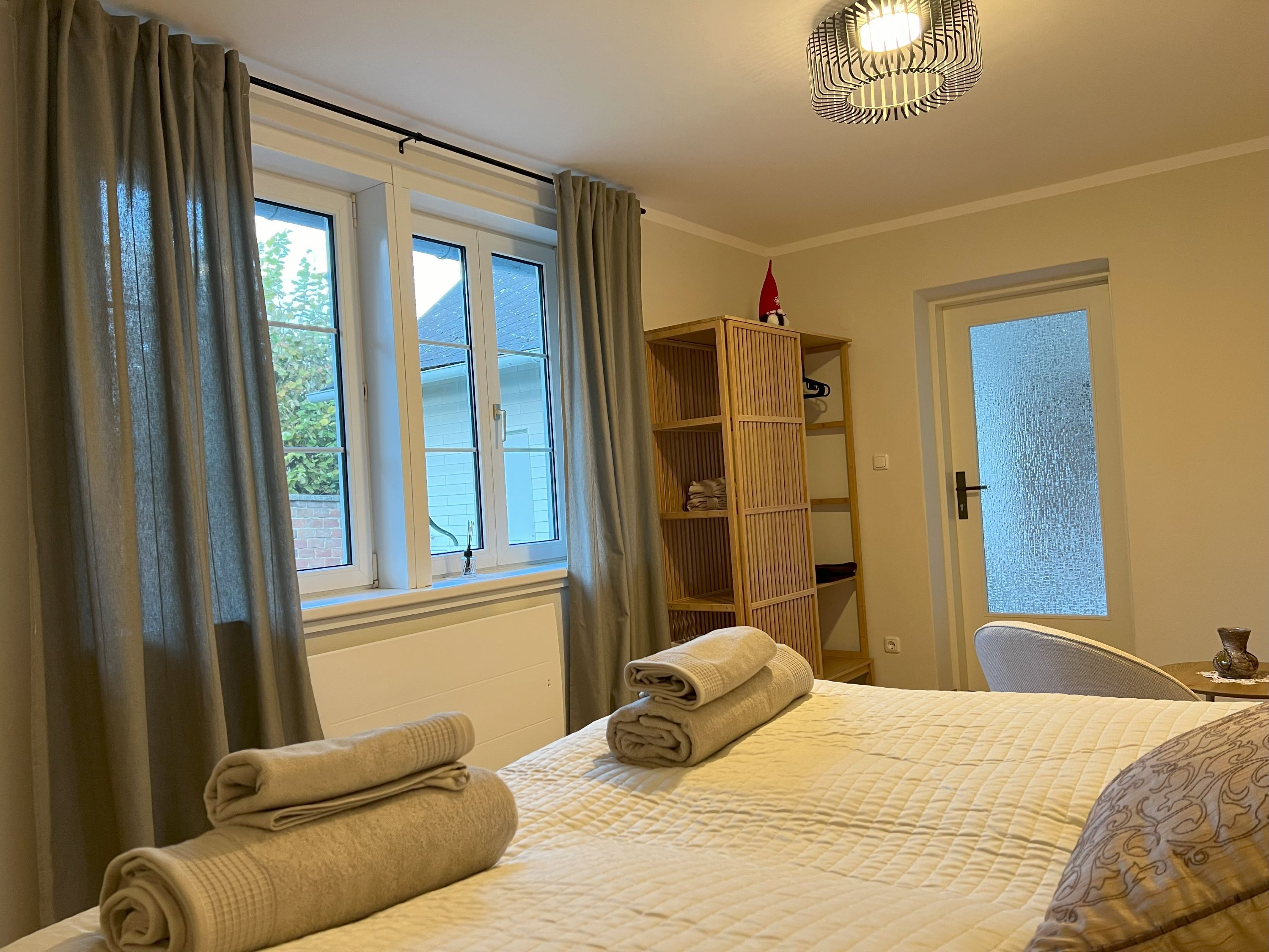 Hangulatos hálószoba, ágy, rajta törölközőkkel, ablak függönnyel és egy polc díszekkel.