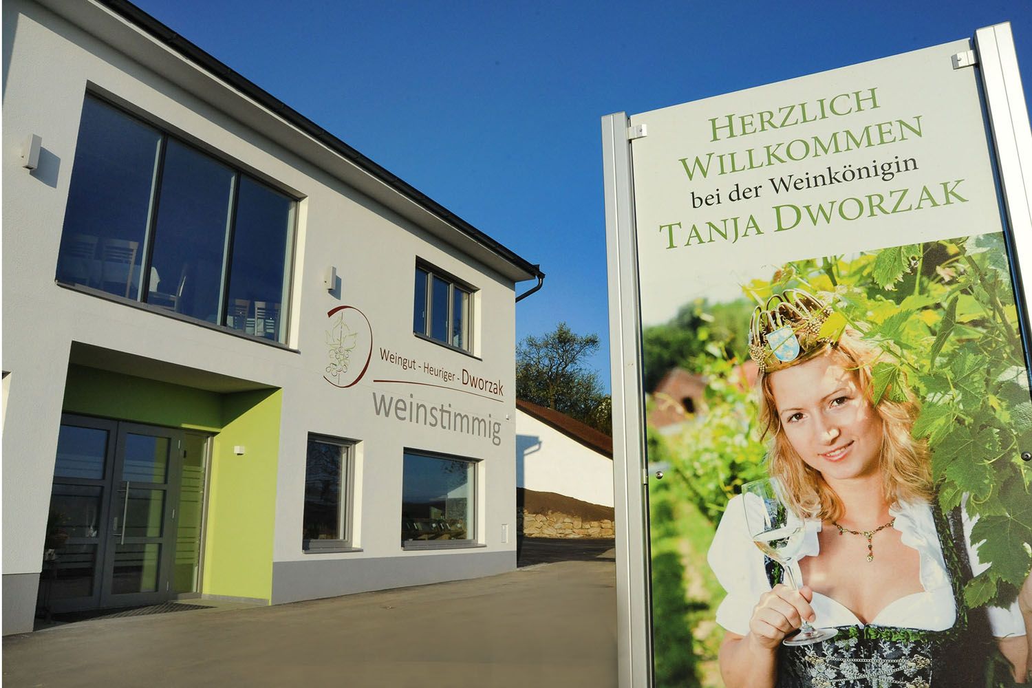 Egy modern épület, amelyen egy felirat látható, amelyen egy népviseletbe öltözött nő látható borospohárral. Szöveg: Tanja Dworzak borkirálynő üdvözöljük.
