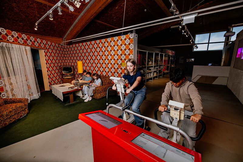 Fiatalok kerékpár ergométeren egy retro nappali dekorációval berendezett helyiségben.